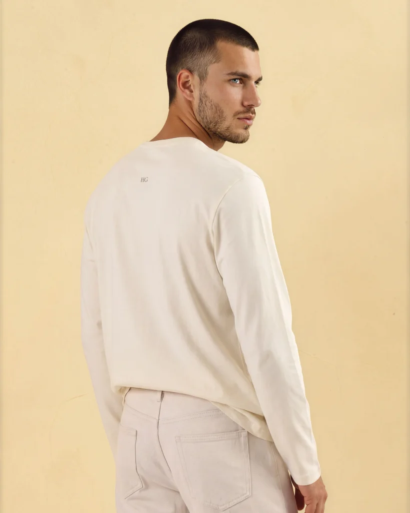Man in a cream Holvo long sleeve top with the HG monogram on the back, looking over his shoulder