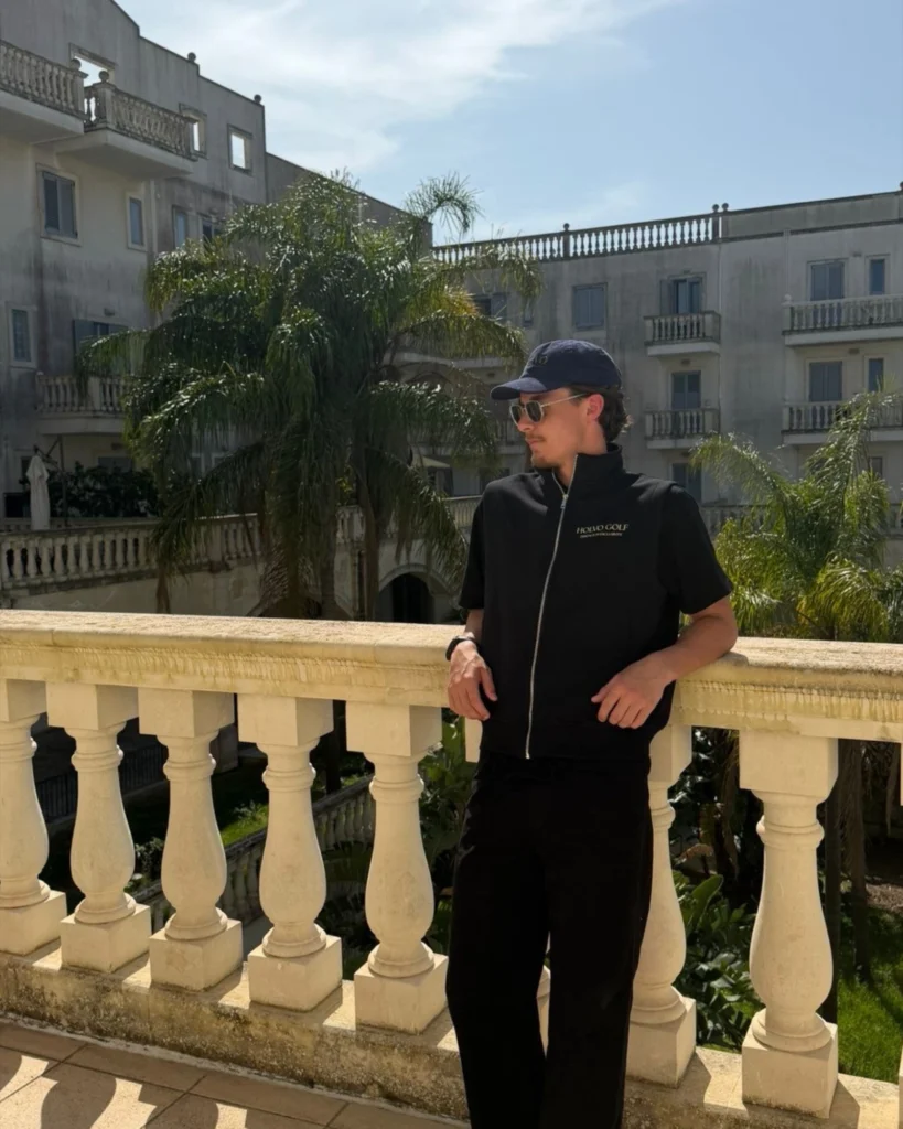 Man in a black short-sleeve Holvo Golf zip top and navy cap, leaning on a stone balustrade