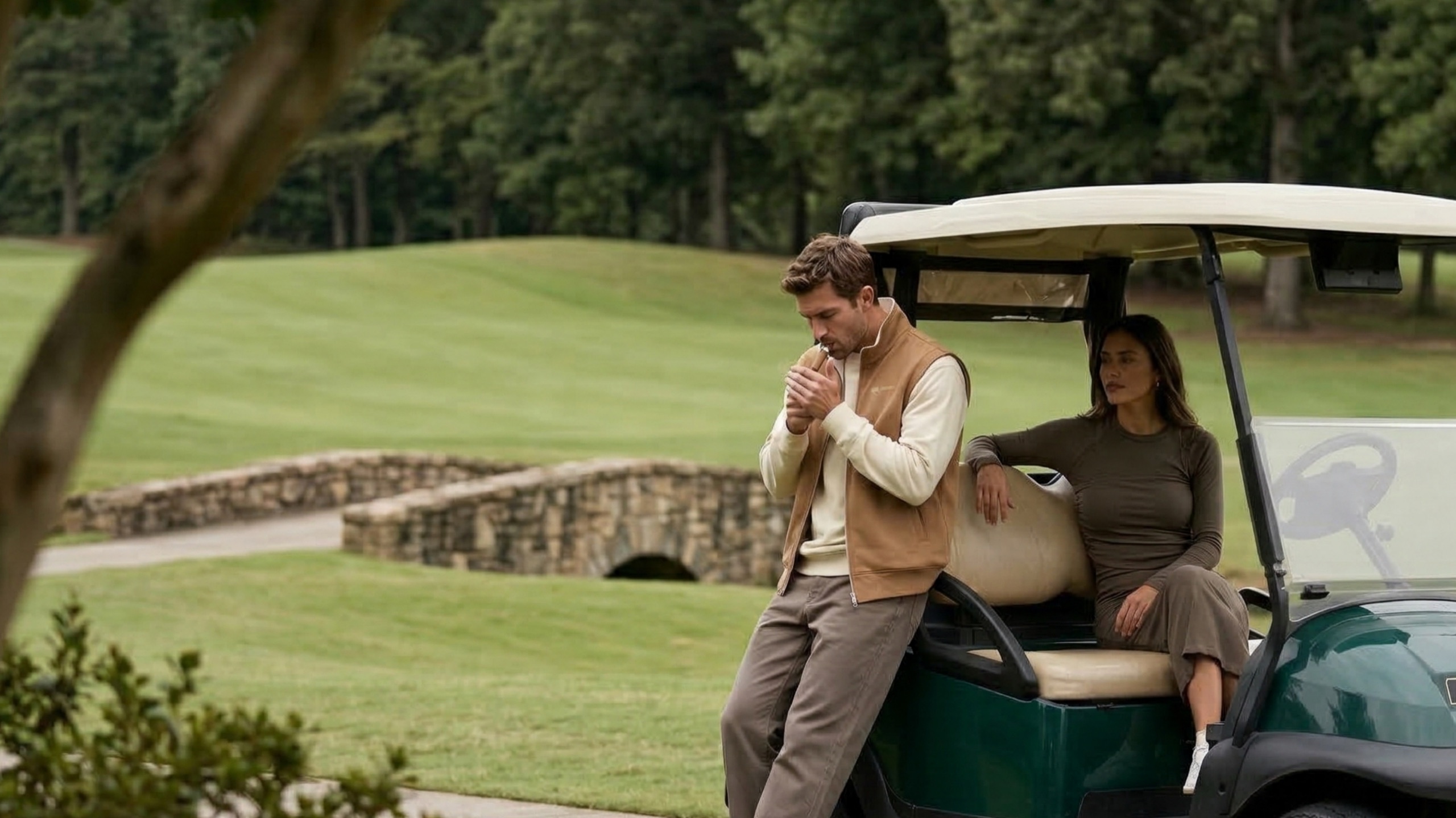 Man in the Holvo Summer Gilet leaning on a golf cart beside a stone bridge, a woman seated in the cart