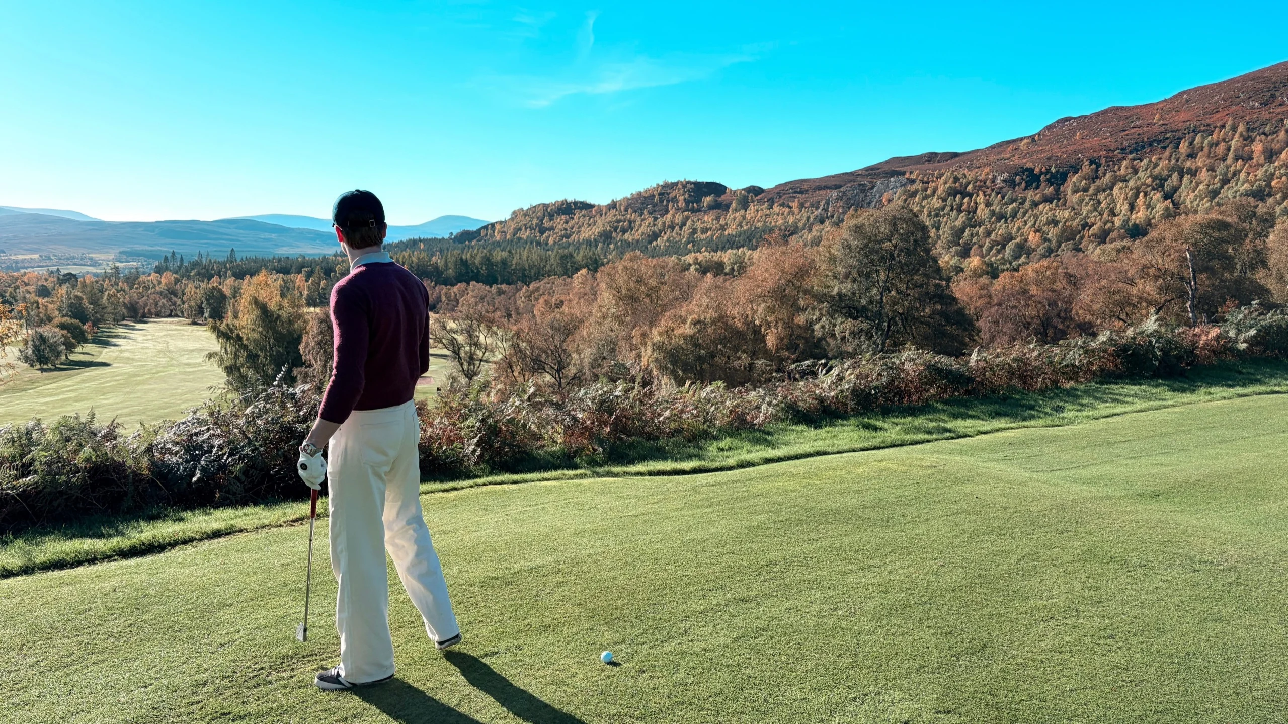 Golfer in a burgundy jumper and cream trousers on the tee, looking out over autumn hills